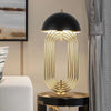 A modern table lamp with a black dome-shaped shade and a gold structure with multiple curved arcs. The lamp is placed on a dark surface, possibly a table, in a room with a light-colored wall and a gray curtain.